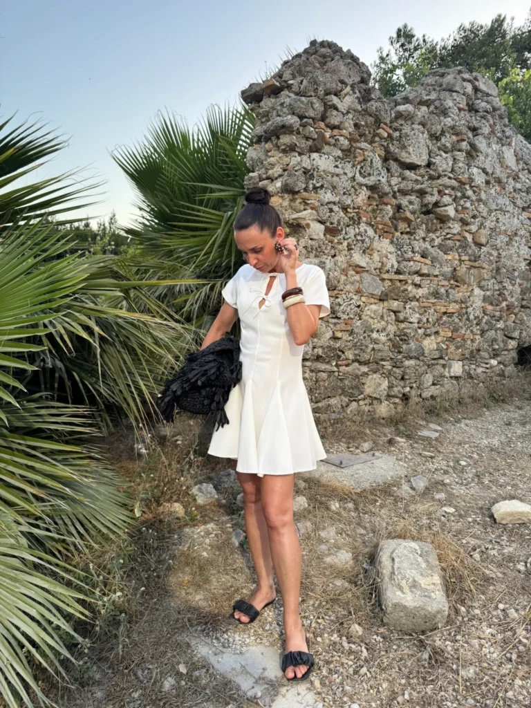 Modne sukienki na lato: Ania Stylistka ubrana w krótką, białą sukienkę i czarne, płaskie sandały pozuje na tle kamiennych ruin i liści palmy.