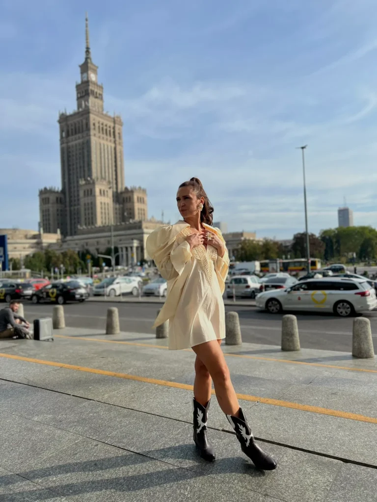 Stylizacja uliczna (street style) w Warszawie: Ania Śmiechowska w kremowej sukience z bufiastymi rękawami i kowbojkach pozuje na tle Pałacu Kultury i Nauki.