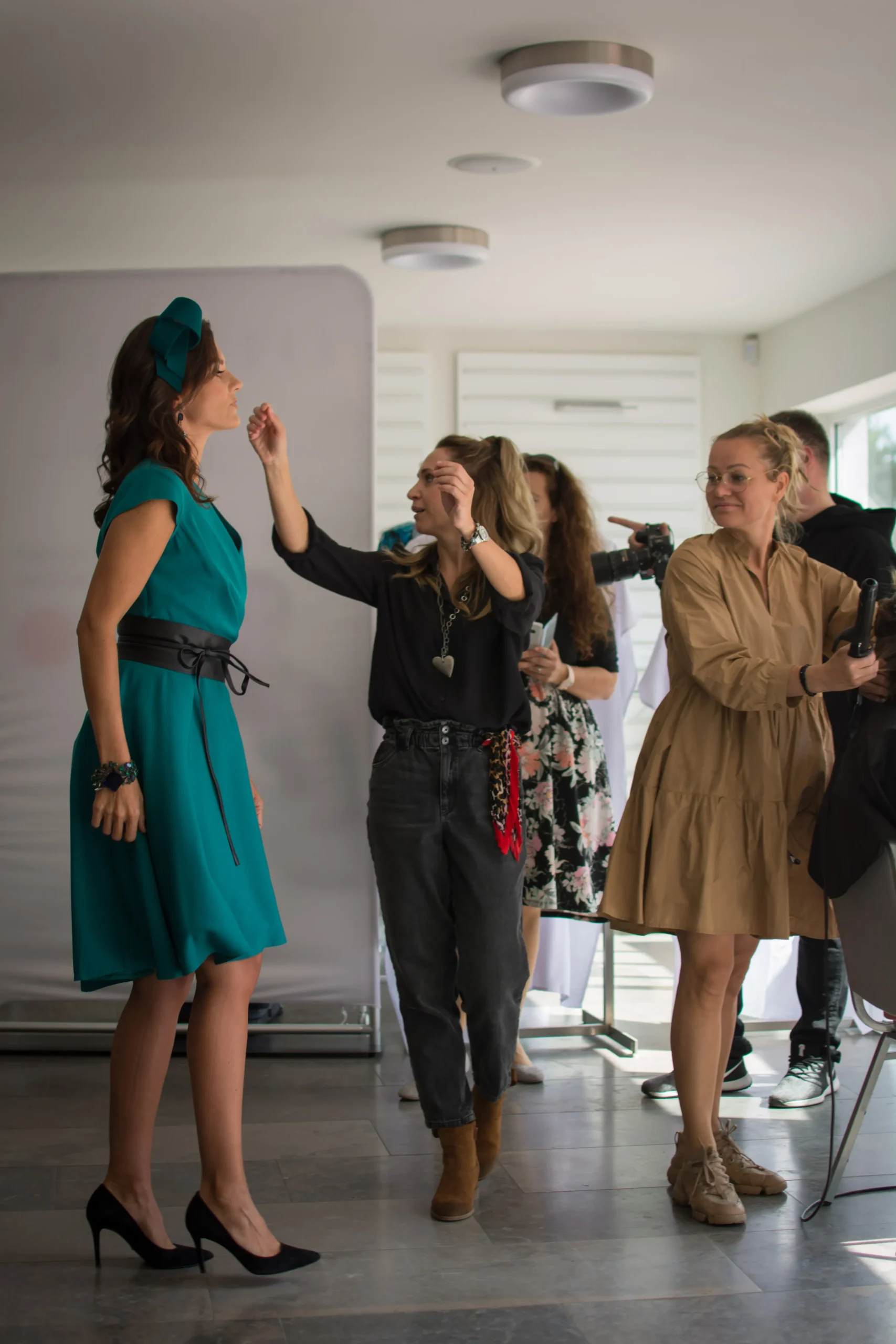 Projekt TyJaMy Zaplecze sesji zdjęciowej. Wizażystka maluje modelkę w zielonej sukience, a w tle fryzjerka układa włosy innej kobiecie.