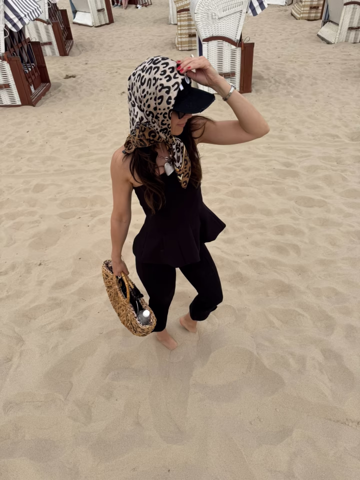 Strona Główna Stylistka mody Ania Śmiechowska w letniej, wakacyjnej stylizacji na plaży. Ma na sobie czarny, elegancki komplet z baskinką, pleciony koszyk oraz oryginalnie zawiązaną chustę w zwierzęcy wzór na czapce z daszkiem.